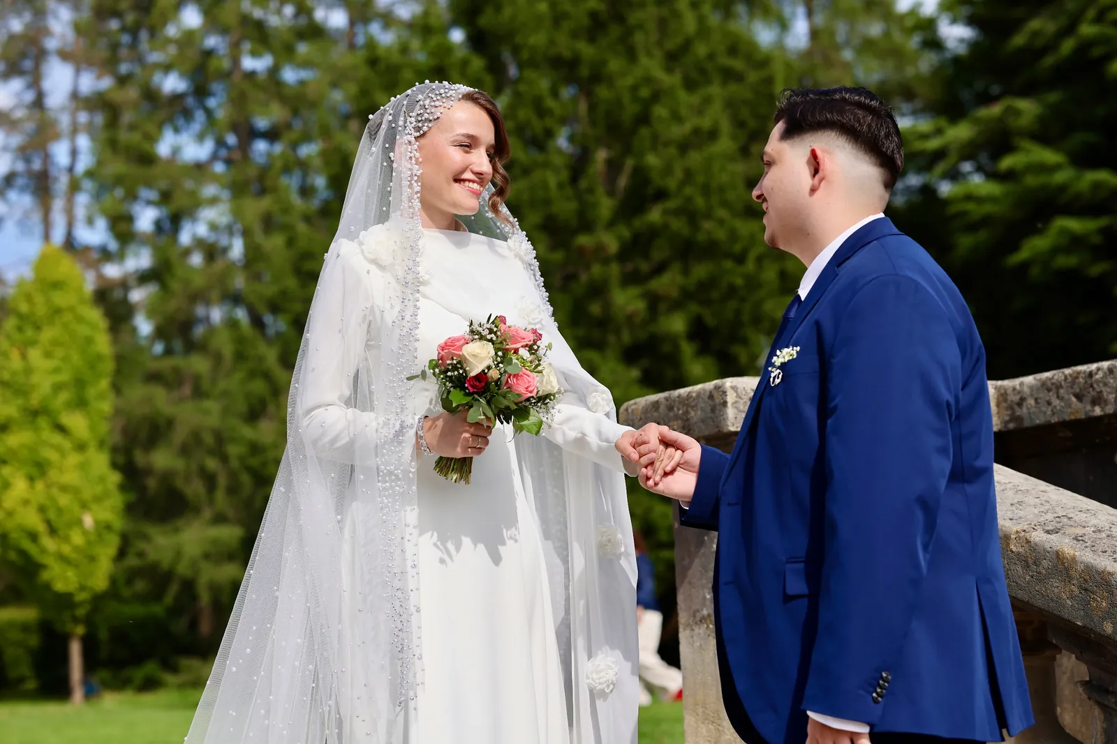 Brautpaar hält sich im Park an den Händen, die Braut mit Perlenschleier und Blumenstrauss