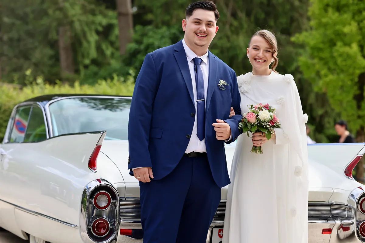 Brautpaar Arm in Arm vor einem weissen Oldtimer mit Heckflossen